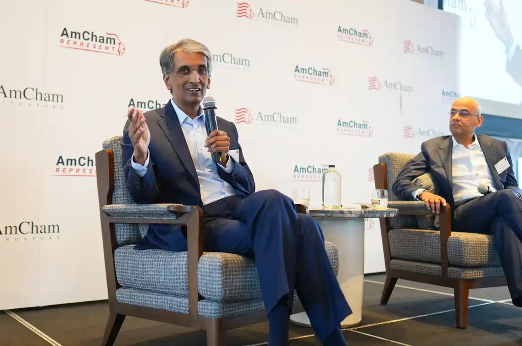 Two speakers in armchairs in front of an AmCham Singapore backdrop
